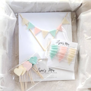 May include: A set of felt cake toppers in a white box. The set includes a felt bunting with pastel green, pink, and cream flags, and a set of felt hearts in pastel green, pink, and cream. The packaging includes the text "Lynne's Mix Felt Cake Topper" and "Lynne's Mix Felt Bunting".