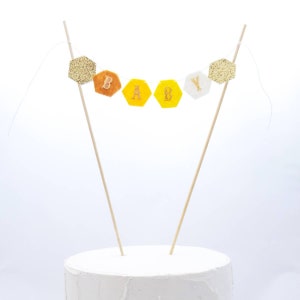 May include: A cake topper with a banner that says "BABY" in gold glitter letters. The banner is made of yellow and orange felt hexagons with gold glitter accents. The banner is attached to two wooden skewers.