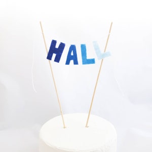 May include: A cake topper with the name "HALL" spelled out in blue felt letters. The letters are attached to wooden skewers and are strung together with white thread.