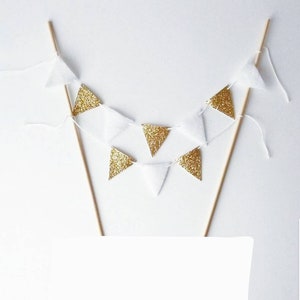 Puede incluir: Una pancarta triangular blanca y dorada con una cadena de triángulos blancos y dorados colgando de dos palos de madera. La pancarta es perfecta para una fiesta o celebración.