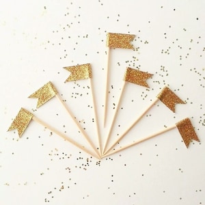 May include: Set of seven gold glitter flags on wooden skewers. The flags are in the shape of a triangle with a pointed tip. The flags are arranged in a fan shape on a white background.