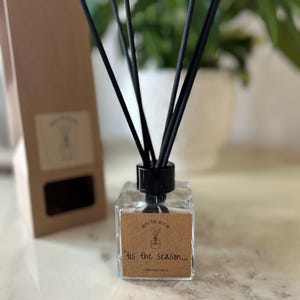 May include: A clear glass bottle diffuser with black reeds and a brown label that reads "Reed the Room 'tis the season... Christmas Spice".