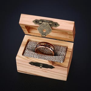 May include: A rose gold ring with a dark inlay and small, sparkling accents sits in an open, light-coloured wooden box. The ring is displayed on a textured, tan fabric. The box has a brass clasp and hinges.