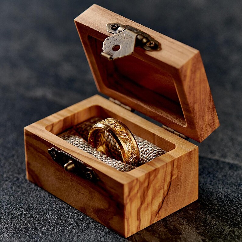 Op de afbeelding: Een gouden ring met een ingewikkeld ontwerp ligt in een open houten kistje. De kist is gemaakt van lichtgekleurd hout en heeft een metalen sluiting. De ring ligt op een textiel van lichte kleur.
