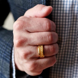 Op de afbeelding: Een goudkleurige ring met een getextureerd oppervlak. De ring wordt aan een vinger gedragen. Het ontwerp van de ring heeft een patroon van kleine, onregelmatige vormen. De achtergrond omvat een blauwe blazer en een geruit overhemd.