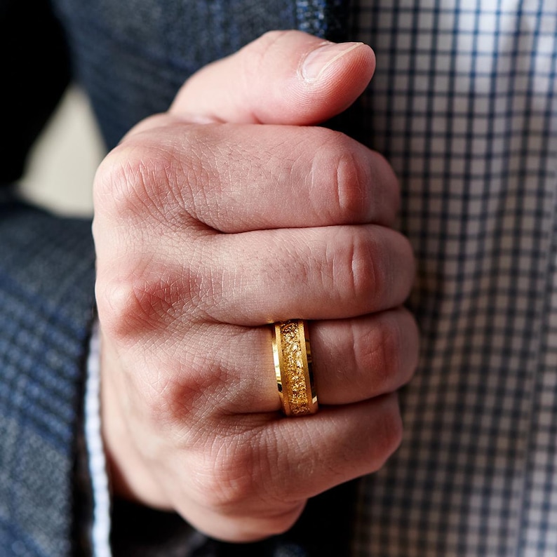 Op de afbeelding: Een goudkleurige ring met een getextureerd oppervlak. De ring wordt aan een vinger gedragen. Het ontwerp van de ring heeft een patroon van kleine, onregelmatige vormen. De achtergrond omvat een blauwe blazer en een geruit overhemd.