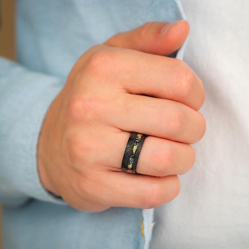 May include: Black ring with a textured pattern and gold-colored accents. The ring is worn on a finger. The ring's design features a dark background with gold-colored leaf-like shapes.