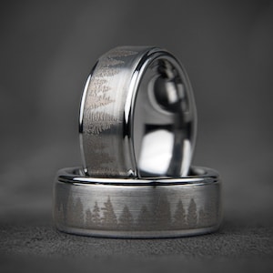 May include: Two silver wedding bands with a brushed finish. The top band has a design of trees etched into the surface. The bottom band has a similar design of trees.