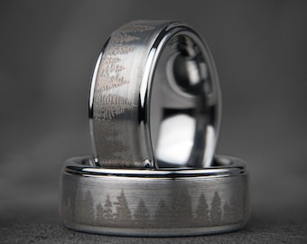 Op de afbeelding: Twee zilveren trouwringen met een geborstelde afwerking. De bovenste ring heeft een ontwerp van bomen dat in het oppervlak is gegraveerd. De onderste ring heeft een vergelijkbaar ontwerp van bomen.