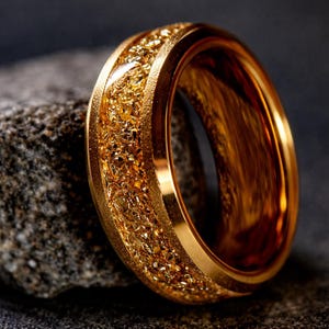 Op de afbeelding: Een goudkleurige ring met een getextureerde, korrelige middenband. De ring staat tegen een donkere achtergrond, wat de metaalglans benadrukt. Het ontwerp van de ring heeft een gladde buitenband en een gedetailleerde, sprankelende binnentextuur.