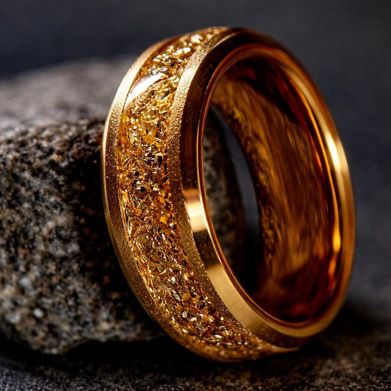 Op de afbeelding: Een goudkleurige ring met een getextureerde, korrelige middenband. De ring staat tegen een donkere achtergrond, wat de metaalglans benadrukt. Het ontwerp van de ring heeft een gladde buitenband en een gedetailleerde, sprankelende binnentextuur.