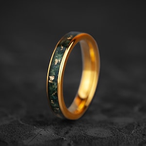 May include: A gold-colored ring with a band of dark green stone inlaid with gold flecks. The ring is set against a dark gray background, highlighting its polished finish and intricate design. The ring's smooth, rounded shape is clearly visible.