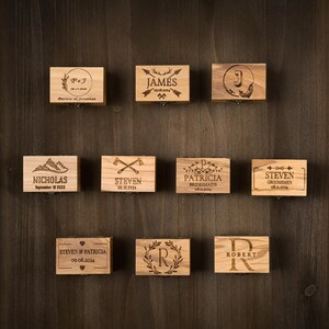 May include: A collection of small, wooden boxes with engraved designs and text. Each box features a different name, initials, or date, with decorative elements like arrows, wreaths, and mountains. The boxes are arranged on a dark wood surface.