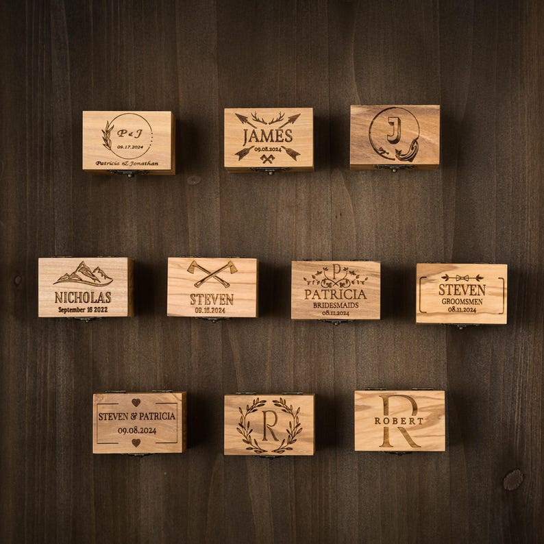 May include: A collection of small, wooden boxes with engraved designs and text. Each box features a different name, initials, or date, with decorative elements like arrows, wreaths, and mountains. The boxes are arranged on a dark wood surface.