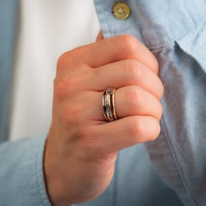 May include: A gold-coloured ring featuring a black and grey inlay and a thin gold band. The ring is on a finger, with a light blue shirt in the background. The ring has a modern, minimalist design.
