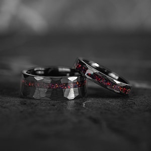 Puede incluir: Dos anillos negros con una textura martillada y una vibrante incrustación roja y púrpura. Un anillo está en posición vertical, mientras que el otro está ligeramente inclinado, mostrando el intrincado diseño. Los anillos están sobre un fondo oscuro y borroso.