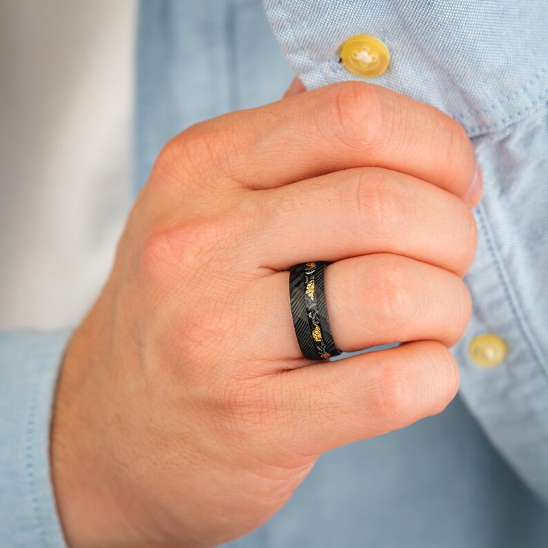 May include: Black ring with gold-colored accents. The ring has a textured surface and is worn on a finger. The ring is a piece of jewelry.