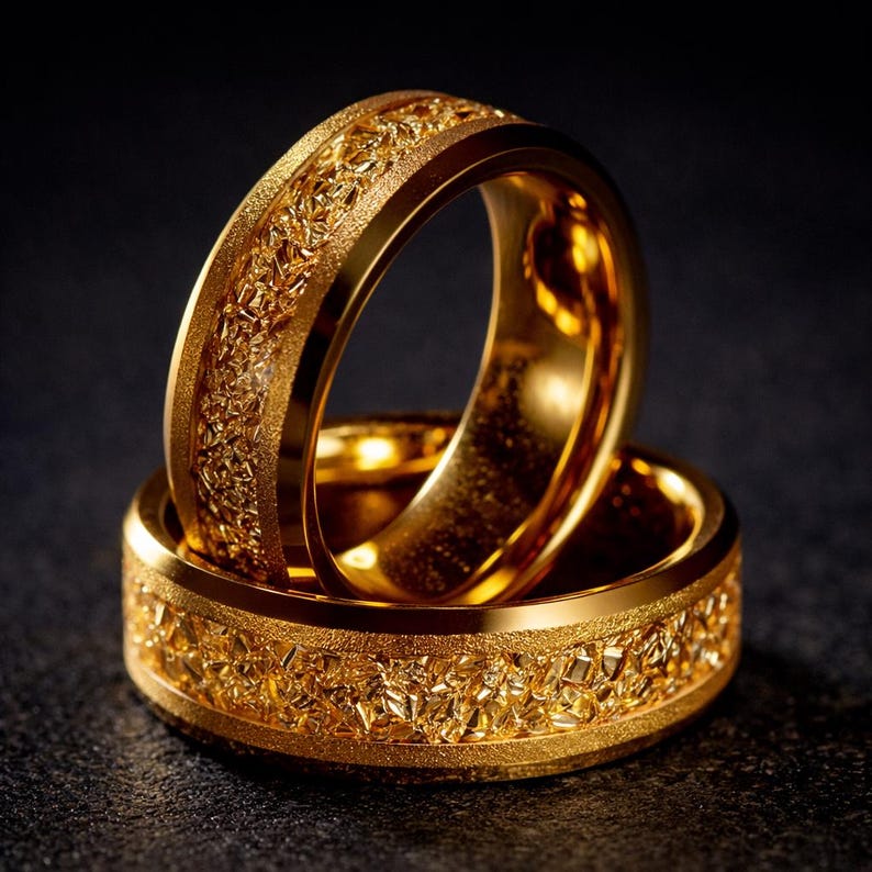 Op de afbeelding: Twee gouden ringen op elkaar gestapeld. De ringen hebben een getextureerd, geometrisch patroon rond de band. De ringen zijn helder, glanzend goudkleurig en de achtergrond is donker.
