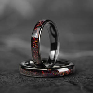 May include: Two matching rings with a dark, polished metal band. The bands are inlaid with a shimmering, speckled material in shades of red, orange, and yellow. The rings are set against a dark, textured background.