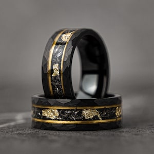 Könnte beinhalten: Zwei schwarze Ringe aus Wolframcarbid mit gehämmerter Oberfläche und einer goldenen Einlage. Die Ringe sind übereinander gestapelt.