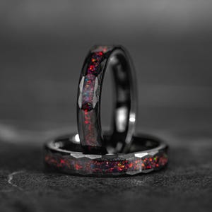 Könnte beinhalten: Zwei schwarze Ringe mit facettiertem Design. Die Ringe haben ein Band aus rotem, orangefarbenem und violettem Opal. Die Ringe sind vor einem dunklen Hintergrund platziert.
