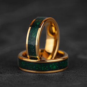 Puede incluir: Dos anillos dorados con una incrustación verde oscuro. Los anillos están apilados, uno ligeramente detrás del otro. Los anillos tienen un acabado pulido y están sobre un fondo gris oscuro.