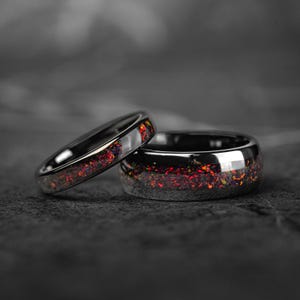 Puede incluir: Dos anillos negros con una vibrante incrustación moteada roja y naranja. Un anillo es más delgado e inclinado, mientras que el otro es más ancho y se mantiene erguido. Los anillos están sobre un fondo oscuro y borroso.