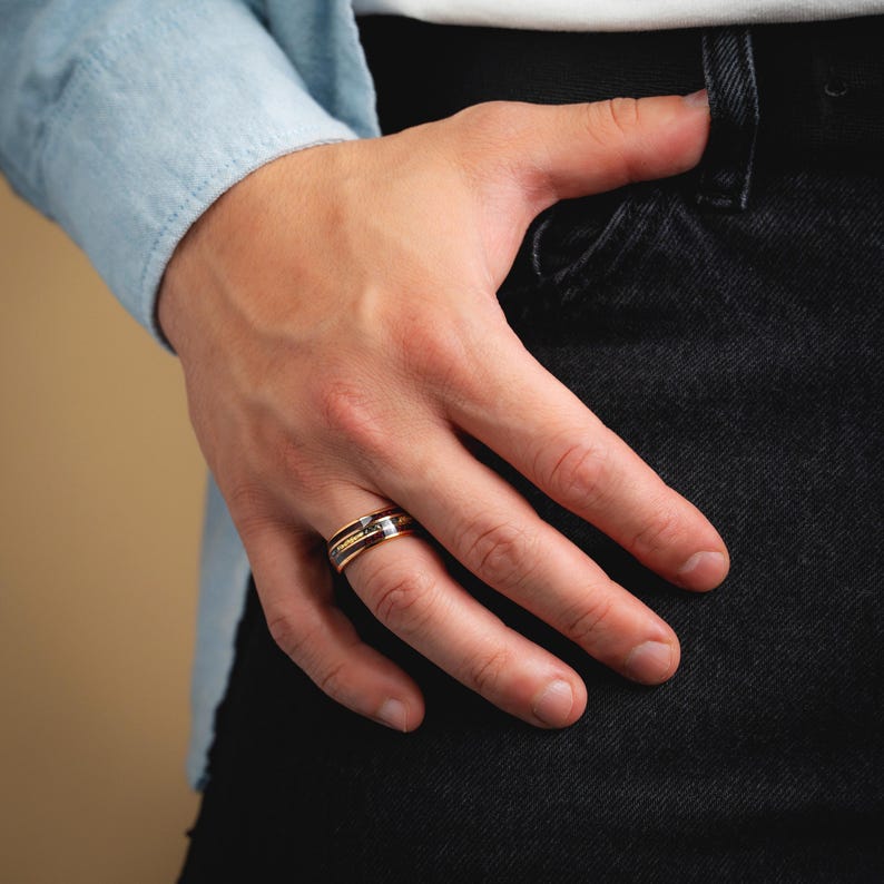 May include: A close-up of a gold, silver, and rose gold ring with a geometric design. The ring is on a person's finger, with a light blue shirt and black jeans in the background. The ring has a small, square-shaped stone.
