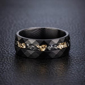 May include: A black ring with a geometric pattern and a gold-colored inlay. The ring has a dark, textured appearance and a band of gold-colored material in the center. The ring is likely made of metal and is designed for wear on a finger.