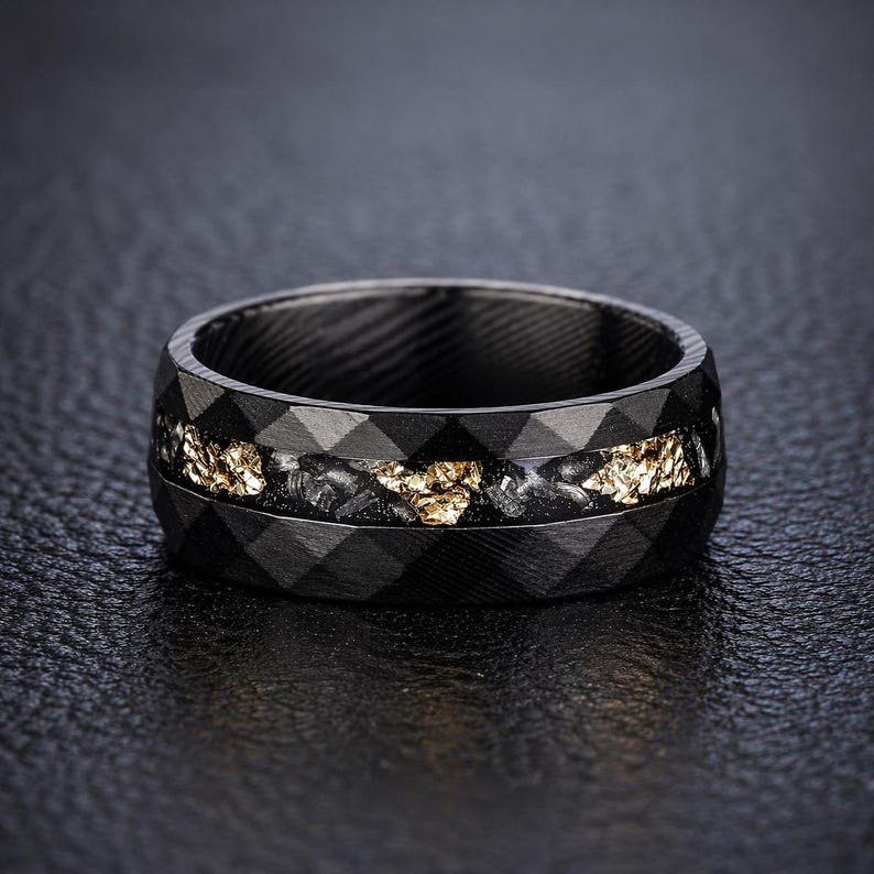 May include: A black ring with a geometric pattern and a gold-colored inlay. The ring has a dark, textured appearance and a band of gold-colored material in the center. The ring is likely made of metal and is designed for wear on a finger.