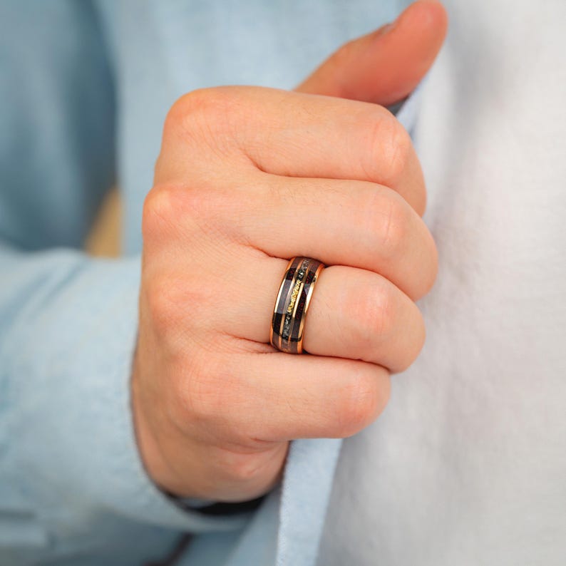 May include: A rose gold ring with black and gold inlays. The ring has a black band with a gold stripe in the centre and gold edges. The ring is worn on a finger.