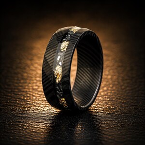May include: A black ring with a carbon fiber pattern and a central inlay of gold leaf and black material. The ring has a wide band and a polished finish, suitable for a wedding or fashion accessory.