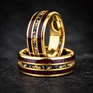 Puede incluir: Dos anillos de tono dorado con una incrustación central de material rojo oscuro triturado, flanqueada por dos bandas doradas finas. Una franja estrecha de motas metálicas doradas y negras recorre el centro de la incrustación roja.