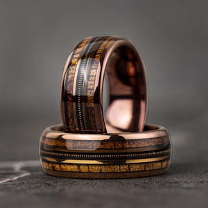 May include: Two copper wedding bands with a wood inlay design. The bands have a dark brown wood inlay with gold accents.