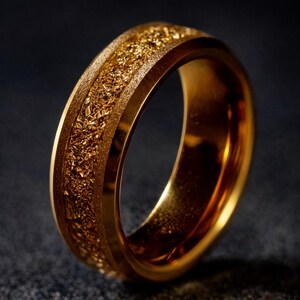 Op de afbeelding: Een goudkleurige ring met een getextureerde middenband. De ring heeft een gladde, gepolijste buitenrand en een ruwe, korrelige textuur in het midden. De ring is gemaakt van een glanzend, metalen materiaal.