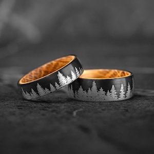 Puede incluir: Dos anillos negros con un diseño de bosque. Los anillos tienen una línea de árboles plateada grabada en el exterior y un interior marrón claro. Los anillos son de metal y se muestran sobre una superficie oscura.
