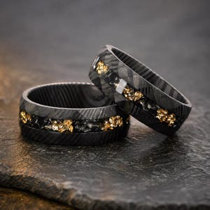 May include: Two black rings with a textured pattern. Each ring features a central band of black stones and gold flakes. The rings are displayed on a dark, textured surface.