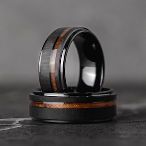 May include: Two black tungsten carbide rings with a brown wood inlay. The rings are stacked on top of each other.