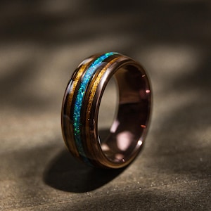 May include: A wooden ring with a central band of blue and green opal. The ring has a dark brown exterior with gold-coloured accents. The ring is set against a dark background.