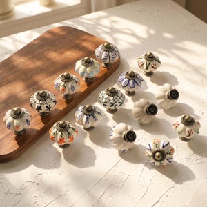 May include: A collection of decorative ceramic cabinet knobs with brass hardware. The knobs feature various floral and geometric patterns in white, blue, orange, and green. They are arranged on a wooden cutting board and a white surface.