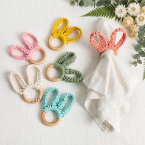 May include: A collection of colorful macrame bunny ear napkin rings in pink, yellow, green, cream, and turquoise. Each ring has a wooden base. One ring holds a white linen napkin. The word "Poppy" is engraved on one ring.
