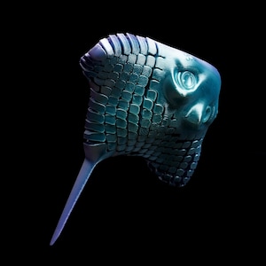 May include: A blue-green, stylized sculpture of a bird's head with a long, pointed beak. The head features a textured, scale-like pattern and a prominent eye. The sculpture is set against a black background.