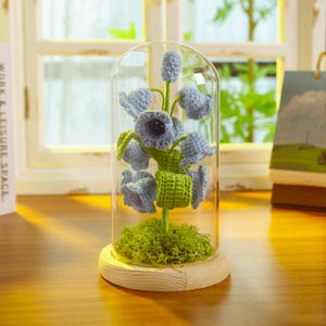 May include: A glass cloche with a wooden base, containing a crocheted flower arrangement. The flowers are blue and green and resemble bells. The cloche is sitting on a wooden table.