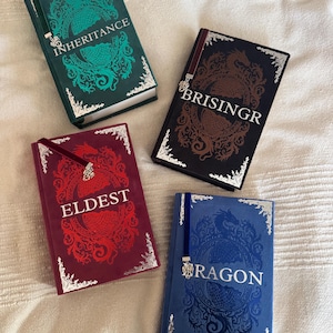 Puede incluir: Se muestran cuatro libros de tapa dura con cubiertas ornamentadas. Los libros son de color verde, negro, rojo y azul. Cada libro tiene un título en letras blancas: "Inheritance", "Brisingr", "Eldest" y "Dragon". Las cubiertas presentan diseños de dragones y florales.