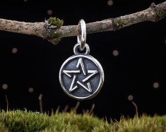 Könnte beinhalten: Ein silberner Anhänger mit einem fünfzackigen Stern, bekannt als Pentagramm, in einem runden Design. Der Anhänger hängt an einem Zweig vor einem schwarzen Hintergrund mit weißen Punkten.