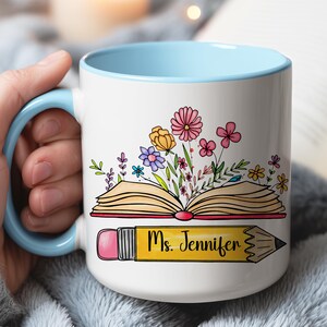 May include: A white ceramic mug with a light blue handle and interior rim. The front features an illustration of an open book with colorful flowers blooming from the pages, positioned above a yellow pencil with the text Ms. Jennifer.