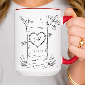 May include: A white ceramic mug with a red handle and interior rim. The exterior features a black line drawing of a tree trunk with a heart carved into the bark. Inside the heart are the letters J+M with an arrow, and the date 08.12.26 below.