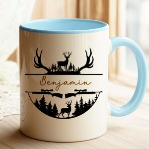 K&ouml;nnte beinhalten: Wei&szlig;er Keramikbecher mit hellblauem Innenraum und Griff. Der Becher zeigt ein schwarzes Silhouette-Design von Hirschen, Gewehren und B&auml;umen, mit dem Namen "Benjamin" in Schreibschrift.