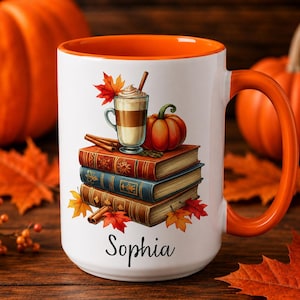 Könnte beinhalten: Ein weißer Keramikbecher mit orangefarbenem Innenraum und Henkel, verziert mit einer Grafik aus drei gestapelten Büchern, einem Kürbis, einer Zimtstange und einem Latte im Glas. Der Name Sophia ist in schwarzer Schreibschrift darunter gedruckt.
