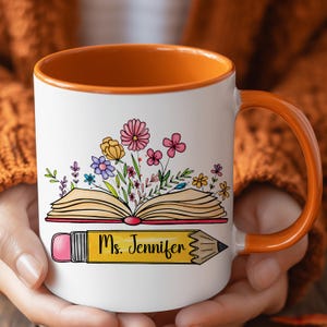 May include: A white ceramic mug with an orange interior and handle. The front features a graphic of an open book with colorful flowers blooming from the pages, positioned above a yellow pencil with the text Ms. Jennifer printed on it.